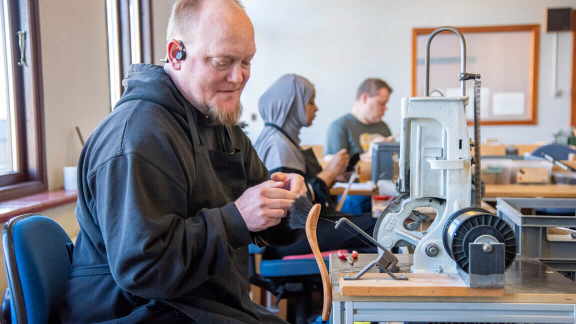 Billedet viser Marc, der sidder ned på en stol og er i gang med at lave en opvaskebørste. I baggrunden sidder der en kvinde og en mand der også laver håndarbejde.