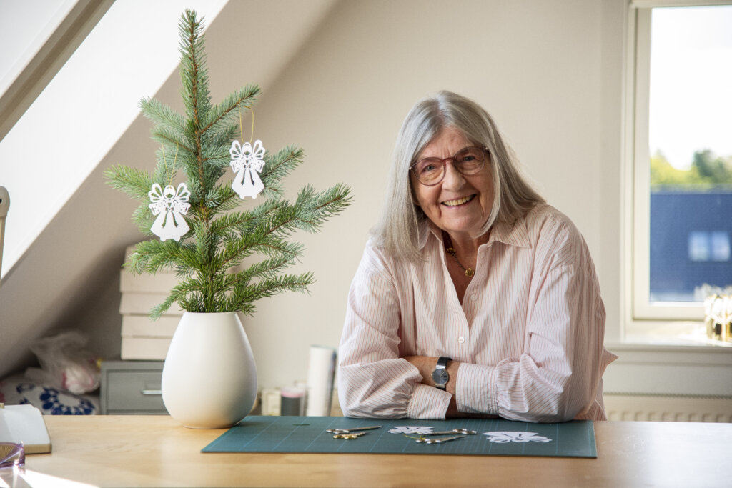 Portræt af smilende Gitte, der sidder ved sit skrivebord, med en grangren ved siden af med julepynten hængende - En papirengel.