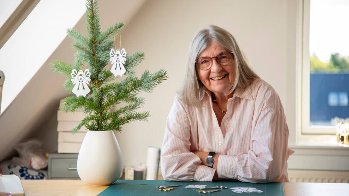 Portræt af smilende Gitte, der sidder ved sit skrivebord, med en grangren ved siden af med julepynten hængende - En papirengel.