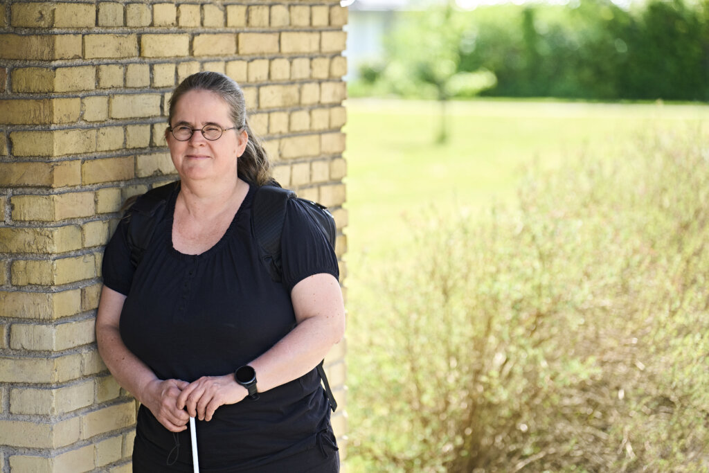 Portræt af 48-årige Karina, som læner sig op ad en mur uden for. Hun holder begge hænder om sin blindestok