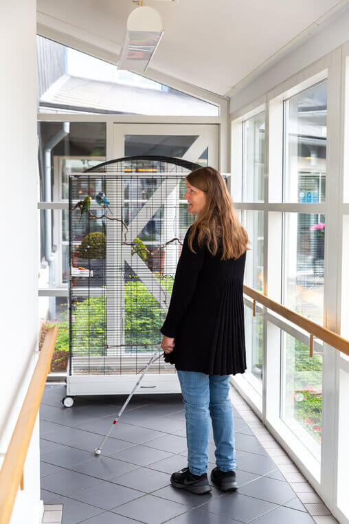 Kvinde med hvid stok st&aring;r i en gang med glasv&aelig;gge og r&aelig;kv&aelig;rk. Foran hende ses et stort fuglebur, som fungerer som orienteringspunkt i rummet.