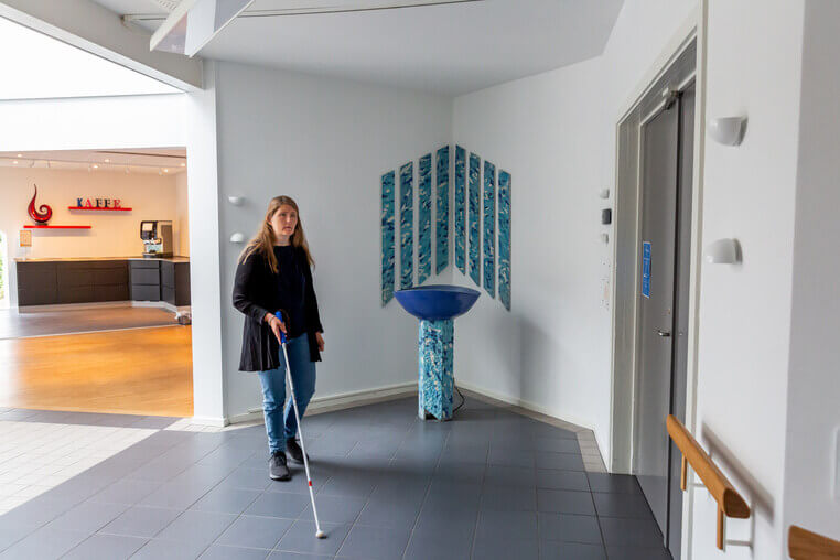 Kvinde med hvid stok st&aring;r i en foyer ved elevator og caf&eacute;omr&aring;de. Taktile v&aelig;gmarkeringer og gulvbel&aelig;gning underst&oslash;tter orientering i rummet.