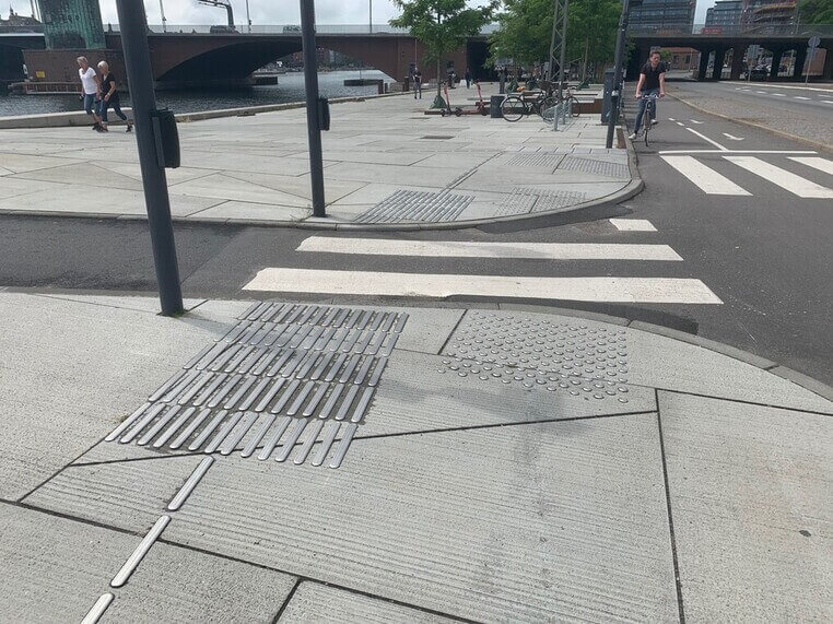 Fodg&aelig;ngerfelt ved vejkryds med taktile ledelinjer og opm&aelig;rksomhedsfelter i bel&aelig;gningen, der guider blinde og svagsynede sikkert over vejen.