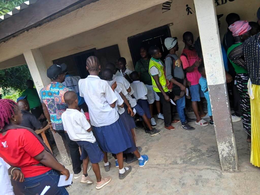 Billedet viser en masse mennesker - både børn, unge & ældre. De står alle sammen i kø udenfor langs en bygning. 