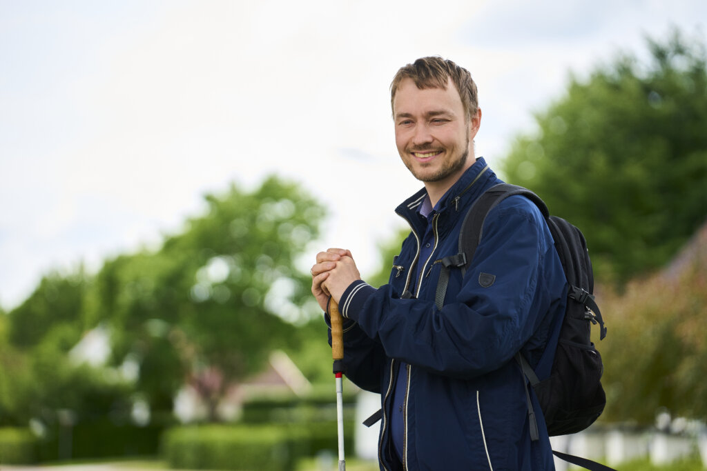 S&oslash;ren smiler ind i kameraet med sin blindestok i h&aelig;nderne. Han er omgivet af tr&aelig;er og natur. 