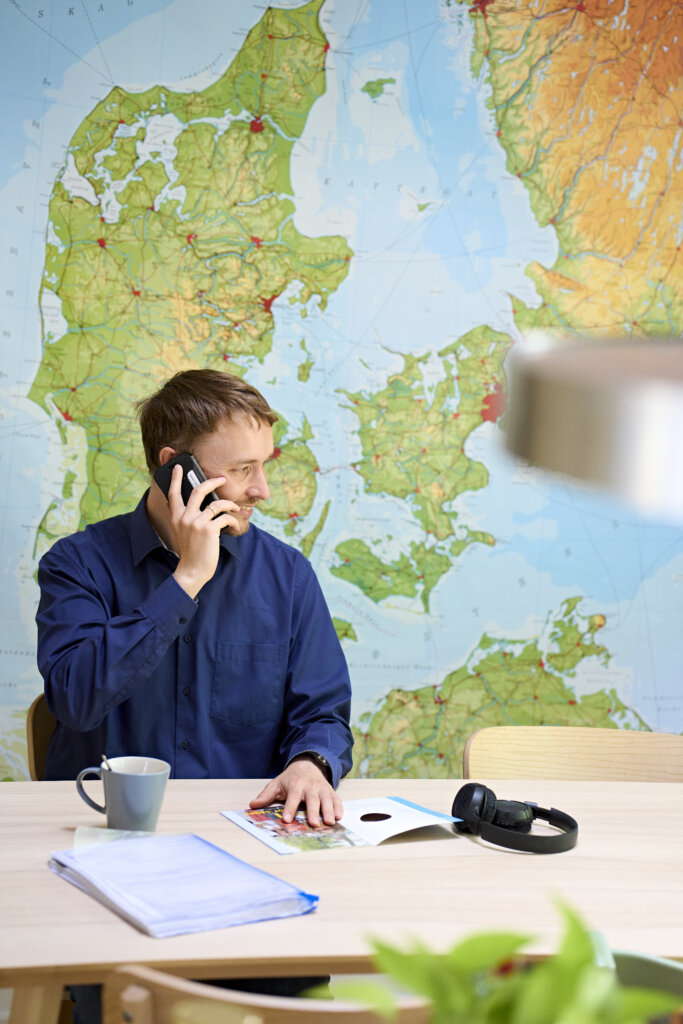 S&oslash;ren sidder ved et skrivebord og snakker i telefon. I baggrunden er der et stort verdenskort. 