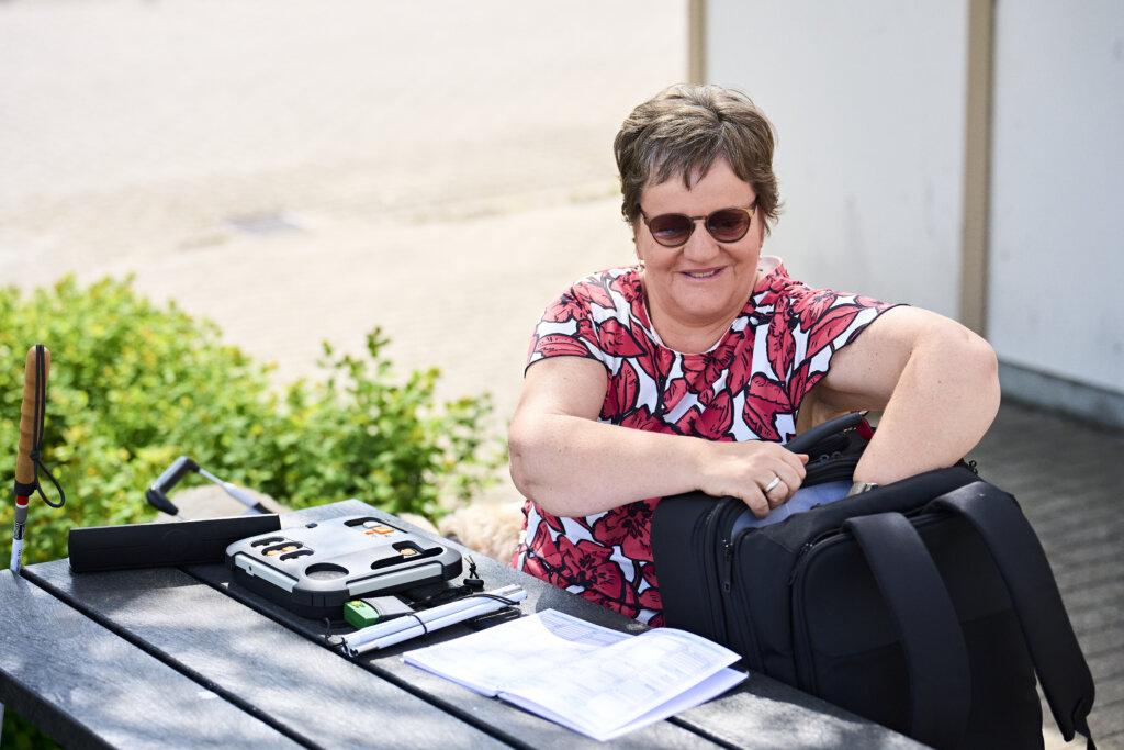Billede af Tina, der sidder p&aring; en b&aelig;nk og smiler mens hun leder efter noget i sin rygs&aelig;k.