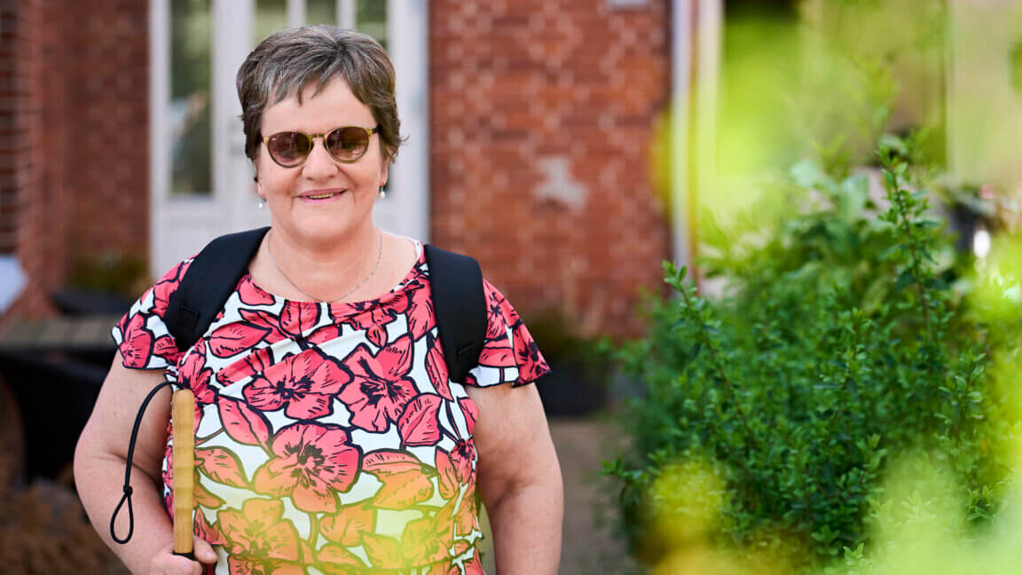 På billedet ser man smilende Tina stå foran et hus, med en rygsæk og solbriller på.