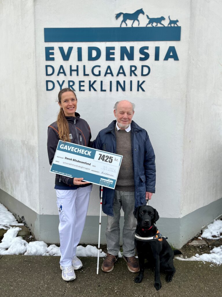 Maria og f&oslash;rerhundebrugeren Kurt st&aring;r foran et skilt, hvor der st&aring;r Evidensia Dahlgaard Dyreklinik. Maria overr&aelig;kker et diplom til kurt. Ved siden af sidder f&oslash;rerhunden Jolly, en sort labrador med f&oslash;rerhundeb&oslash;jle