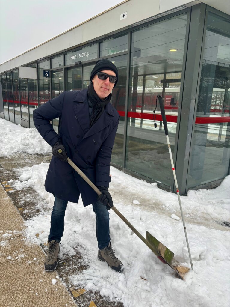 Christian Bundgaard, der er blind, st&aring;r p&aring; en togperron og skovler sne