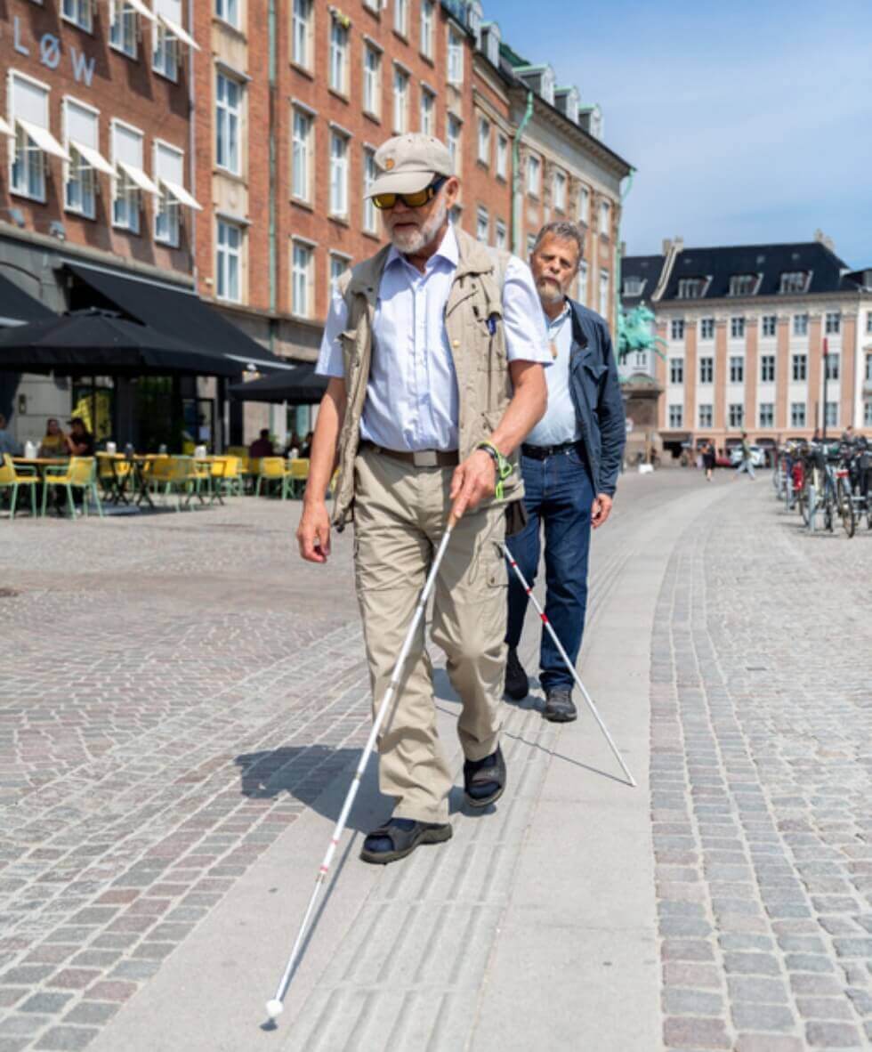 To mænd går langs en taktil ledelinje i brostensbelagt gade ved Gammel Strand. Begge bruger hvid stok til at orientere sig.