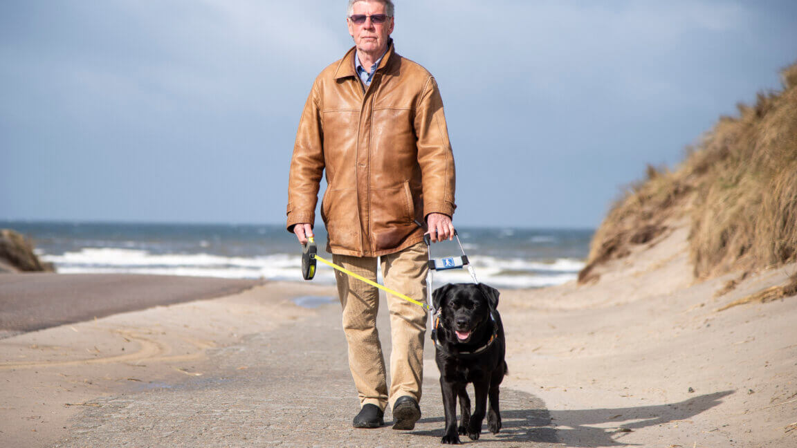Hans Verner kommer gående på stranden med førerhunden Siffi i bøjle, en sort labrador