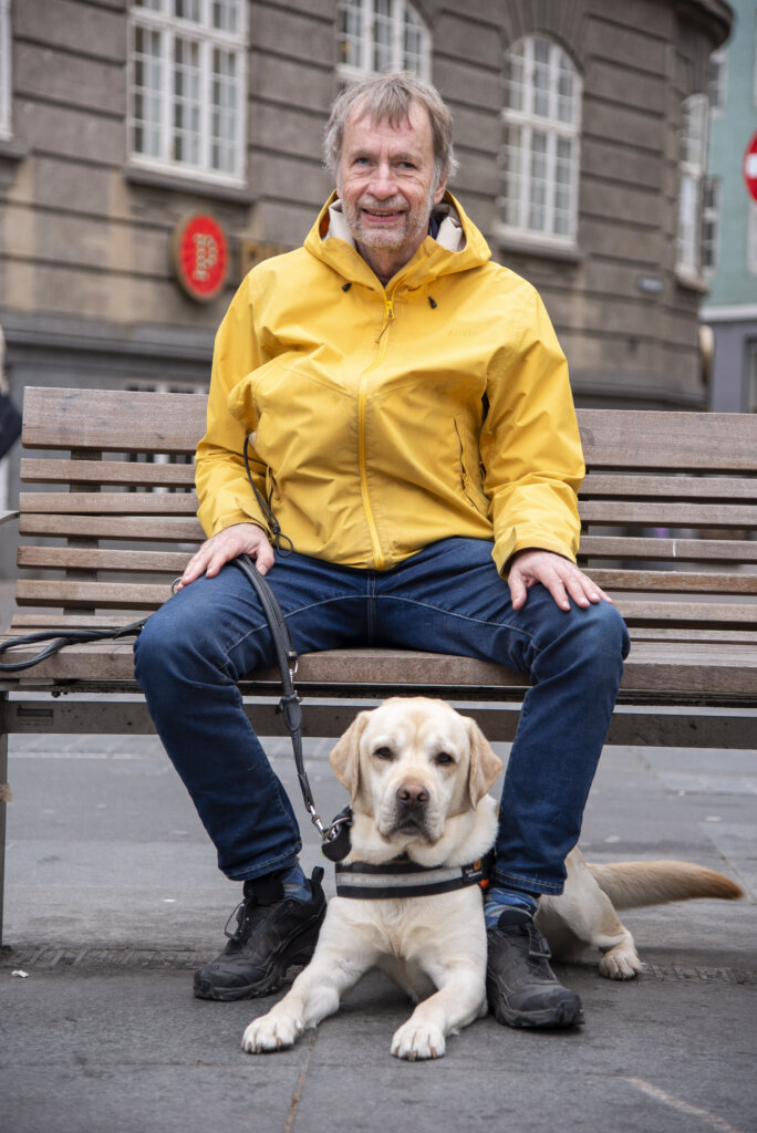 Michael sidder på en bænk ved Nørreport station med førerhunden Pluto mellem benene, en lys labrador med førerhundebøjle