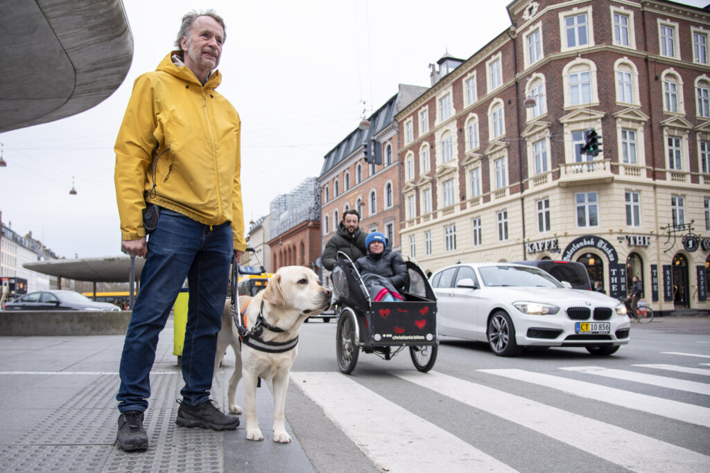 Michael og førerhunden Pluto, lys labrador med førerhundebøjle, venter ved en fodgængerovergang ved Nørreport station