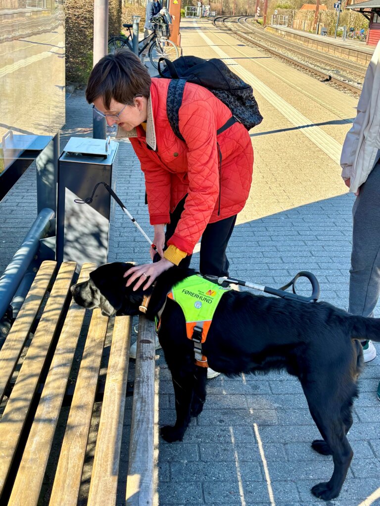 Aston lægger hovedet på en bænk på en tog station. Lene står ved siden af