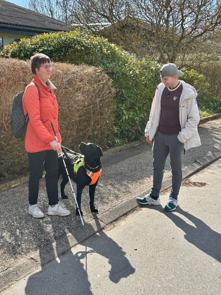Førerhunden Aston, en sort labrador, stopper ved en kantsten og kigger på Lene. Ved siden af står Morten