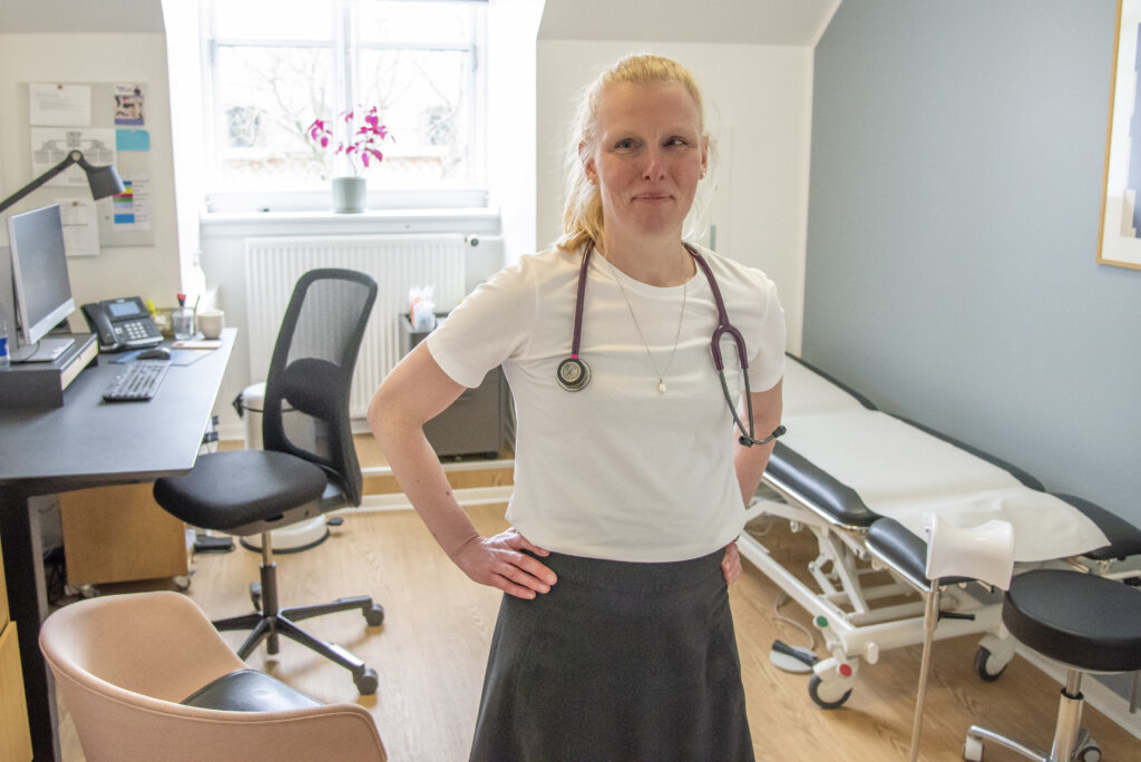 Stine st&aring;r i et konsultationsrum med stetoskop om halsen. I baggrunden st&aring;r et skrivebord og en briks