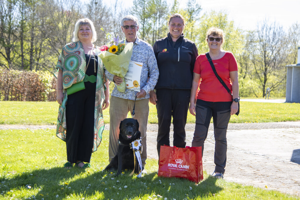 Hans Verner holder en buket blomster og et gavekort. Ved siden af ham sidder f&oslash;rerhunden Siffi, en sort labrador og ved siden af st&aring;r Siffis opdr&aelig;tter, foderv&aelig;rt og instrukt&oslash;r