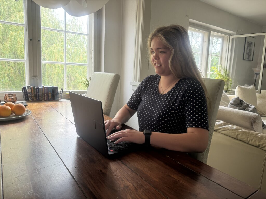 Sarah sidder ved et spisebord og arbejder p&aring; sin computer