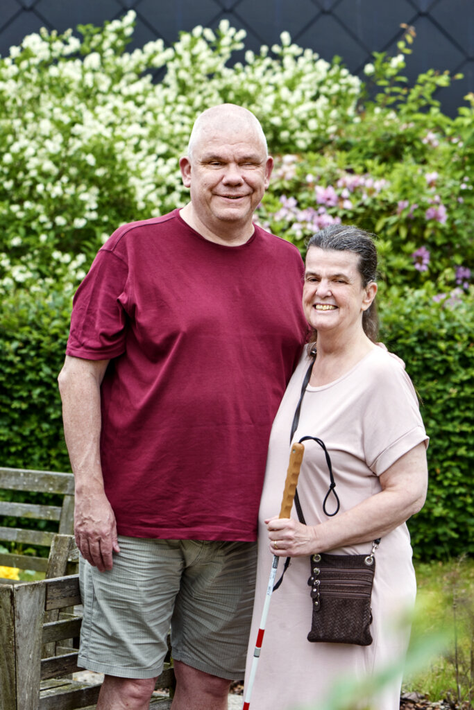 Arne holder om sin kone Joy, der står med en blindestok. De står begge uden for