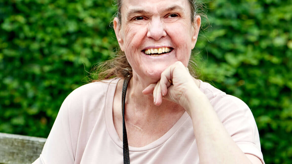 Billede af konsulenten Joy, som smiler til kameraet. Joy sidder på en bænk og sidder blandt grønne omgivelser, med en busk og blomster i baggrunden.