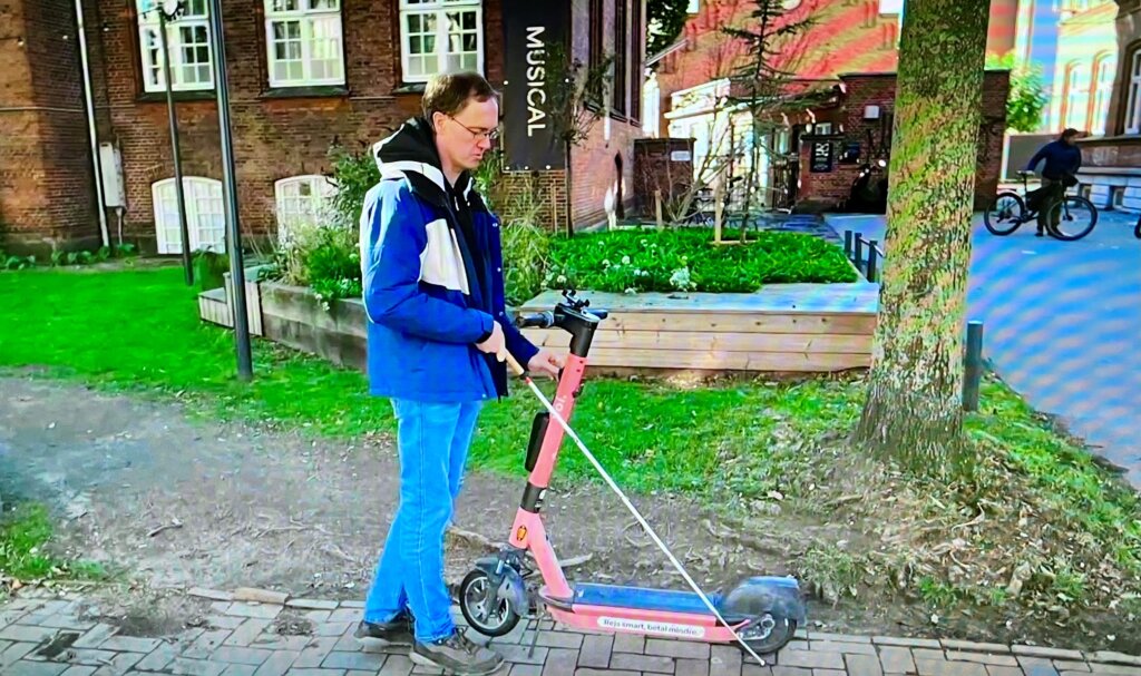 Erik st&aring;r med sin hvide stok foran et ell&oslash;behjul, der er parkeret p&aring; fortovet