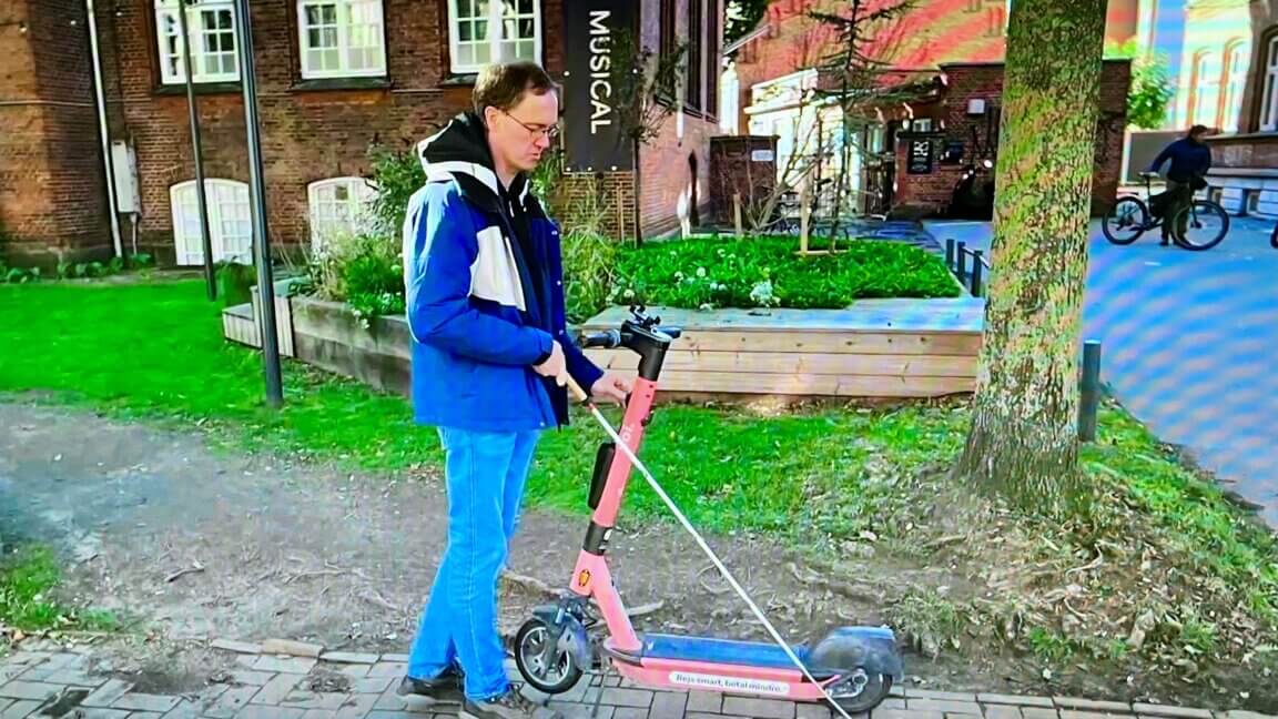 Erik står med sin hvide stok foran et elløbehjul, der er parkeret på fortovet