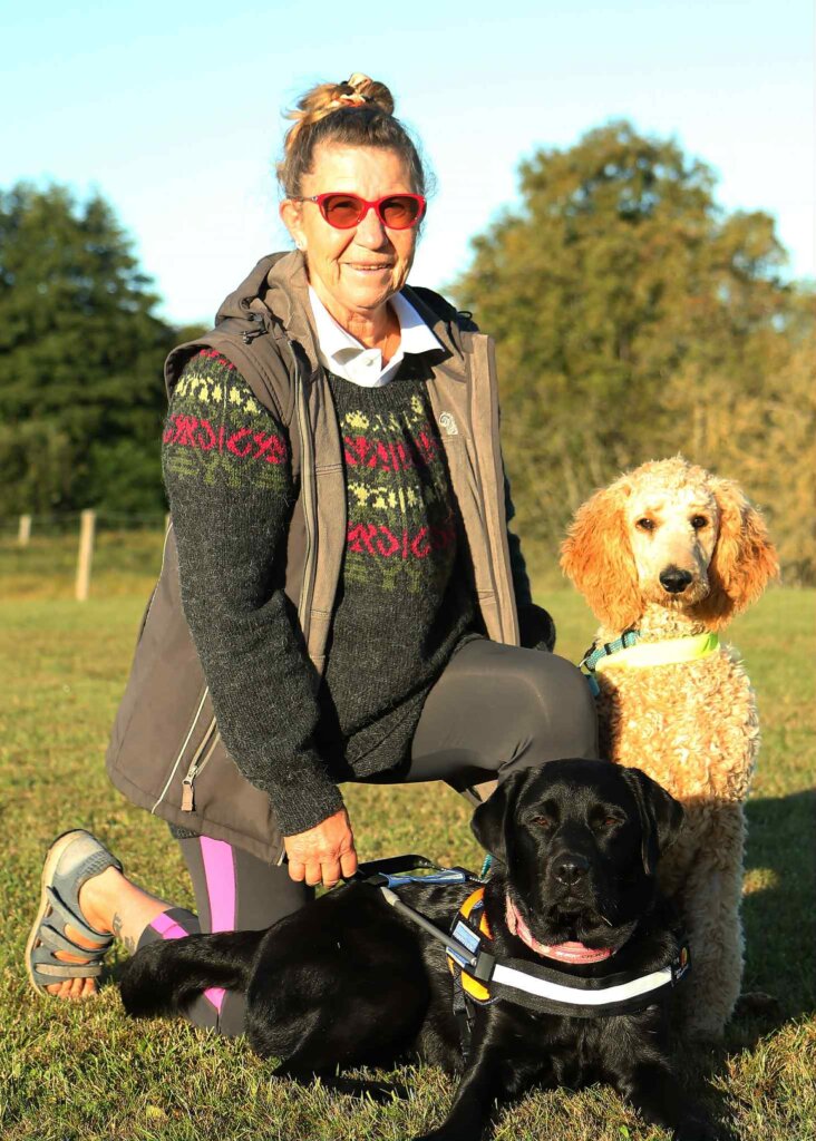 Bente sidder på hug med storpudlen Tell og den sorte labrador Tila, som er i træning til førerhund og derfor iført førerhundebøjle